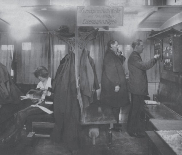 Zwei Männer stehen im Zugabteil vor einem Münzfernsprecher. Einer telefoniert bereits, einer wählt.