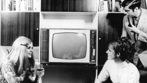 Schwarz-Weißn Fotografie von einem zentral stehenden Fernseher, der von zwei jungen Frauen flankiert wird.