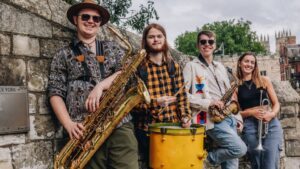 Vier Personen mit Instrumenten, darunter zwei Saxofone, eine Trompete und eine Trommel
