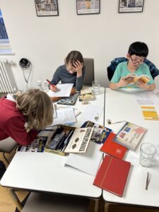 3 Kinder sitzen um einen Tische, der mit Stiften, Sammlerheften und Papier über dem Rätsel aus dem Heft Junge Sammler brüten
